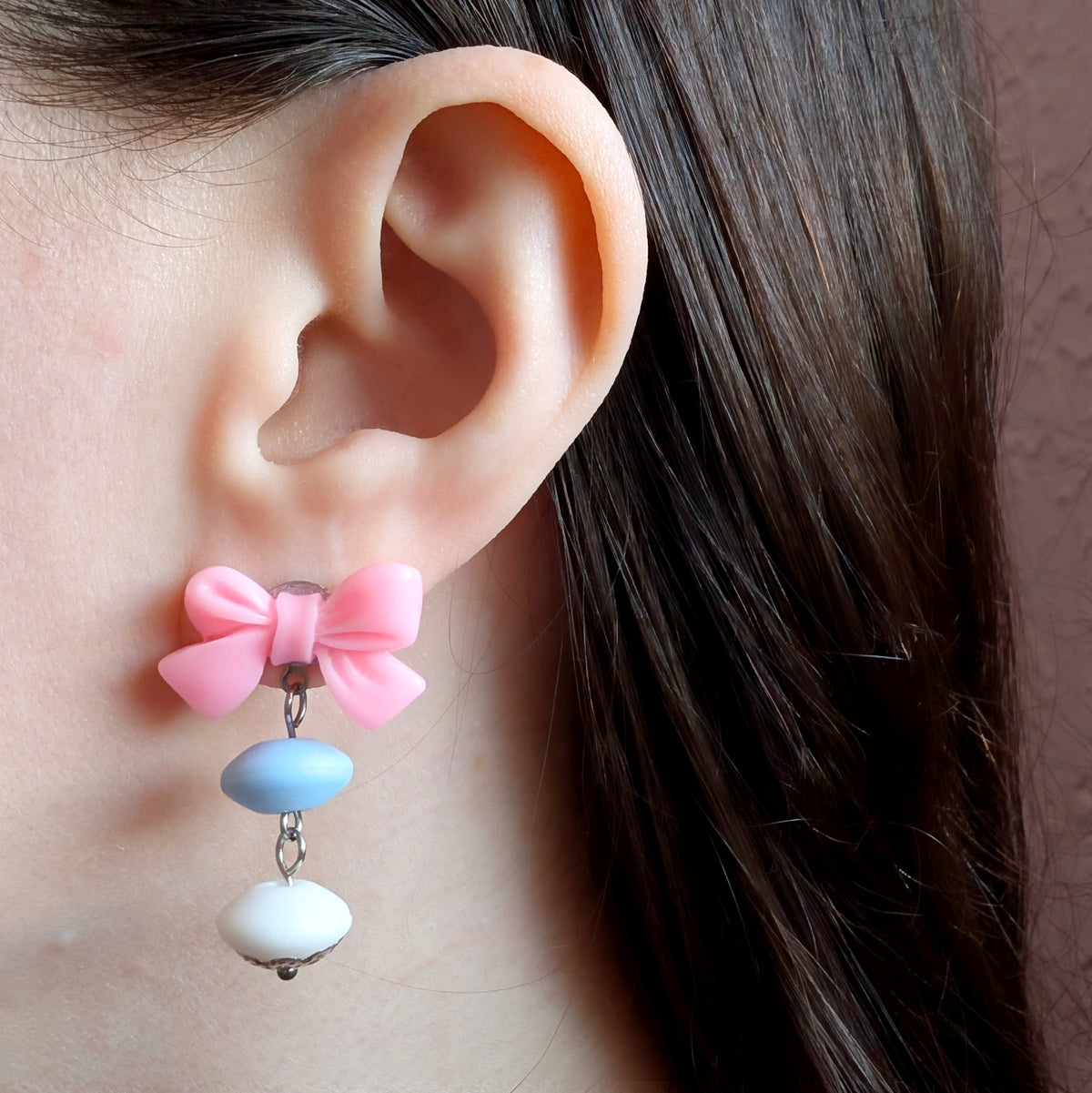 Model photography showcasing the scale and lightweight drape of the asymmetric pastel faux candy earrings with pink resin bow and cascading 12mm matte clay beads.